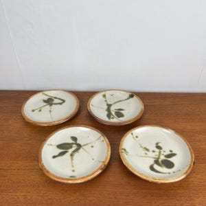 May include: Four small, round ceramic plates with a cream-colored surface and a brown rim. Each plate features a unique abstract design in shades of green and brown. The plates are arranged on a wooden surface.