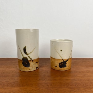 May include: Two ceramic cups with a cream-colored upper half and a tan lower half. Each cup has a unique black and brown abstract design. One cup is taller than the other, both are cylindrical.