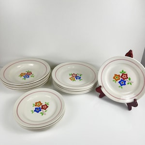 May include: A collection of cream-colored ceramic plates with a red rim and a floral design in the center. The flowers are in shades of orange, red, blue, and yellow. The plates are stacked and displayed on a white surface.