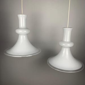 Puede incluir: Dos lámparas colgantes blancas con una pantalla acampanada en forma de campana y un cuerpo redondeado en forma de reloj de arena. Las luces están suspendidas del techo por finos cordones blancos. Las pantallas tienen un sutil borde transparente alrededor del borde inferior.