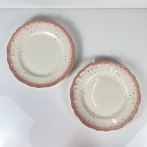 Könnte beinhalten: Zwei weiße Keramikplatten mit gewelltem Rand und rosa Rand. Die Teller haben kleine rosa Tupfen um den Rand. Die Teller stehen auf einer weißen Oberfläche.