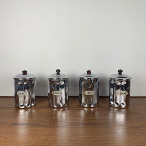 Puede incluir: Cuatro botes de plata con tapas negras y etiquetas que dicen "Farine", "Licorice", "Pates" y "Sucre".