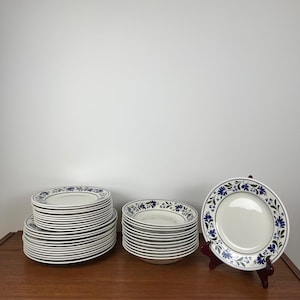 Peut inclure: Une collection d'assiettes en céramique blanche avec un motif floral bleu sur le bord. Les assiettes sont empilées en plusieurs piles sur une surface en bois, avec une assiette exposée sur un présentoir en bois.