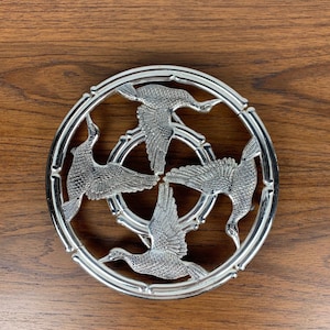 May include: A silver-toned metal hot pad with a circular design featuring four stylised birds in flight. The birds are arranged in a circular pattern, with their wings spread out. The hot pad has a smooth, polished finish.