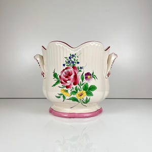 Pode incluir: Um vaso de cerâmica na cor creme com borda recortada e duas alças laterais. O exterior apresenta um motivo floral pintado à mão com uma grande rosa central rosa, flores amarelas menores, flores roxas e folhas verdes na base rosa.