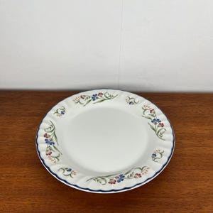 Könnte beinhalten: Weißer Keramikteller mit gewelltem Rand und blauem Rand. Der Teller ist mit einem Blumenmuster mit blauen, roten und grünen Blumen und Blättern verziert. Der Teller steht auf einer Holzoberfläche.