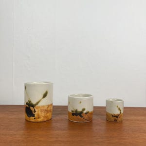 May include: Three ceramic cups of varying sizes. Each cup has a white upper half and a lower half with a textured, earthy brown color. Dark green and black abstract designs decorate the white portion of the cups.