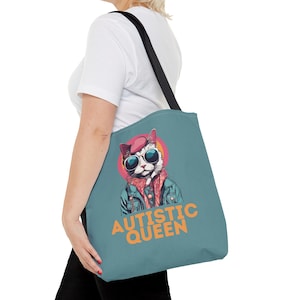 Può includere: Una borsa tote blu-verde con una tracolla nera. La borsa presenta un gatto dei cartoni animati con occhiali da sole e un berretto. Il testo "AUTISTIC QUEEN" è stampato in arancione sulla borsa.
