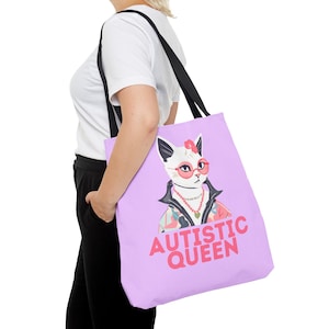 Può includere: Una borsa tote viola con un gatto bianco che indossa occhiali da sole rosa e una camicia floreale. Il testo "AUTISTIC QUEEN" è stampato in rosa sulla borsa.