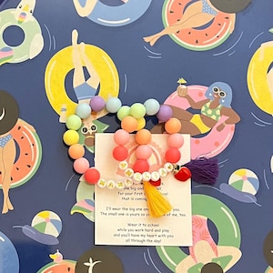 May include: A beaded bracelet with the name "AMELIA" in white letter beads, featuring a rainbow of pastel-coloured beads and a purple tassel. The background is a blue pattern with illustrations of people in inflatable rings.