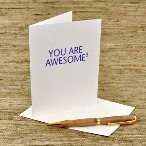 May include: A white card with the text "YOU ARE AWESOME^3" printed in blue ink. A wooden pen with a gold accent is lying on a white envelope.