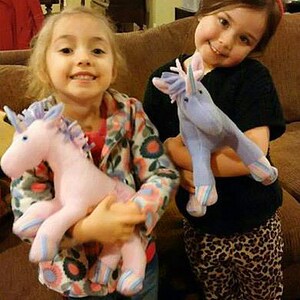 May include: Two young girls are holding plush unicorn toys. One unicorn is pink with blue stripes and the other is purple with blue stripes.