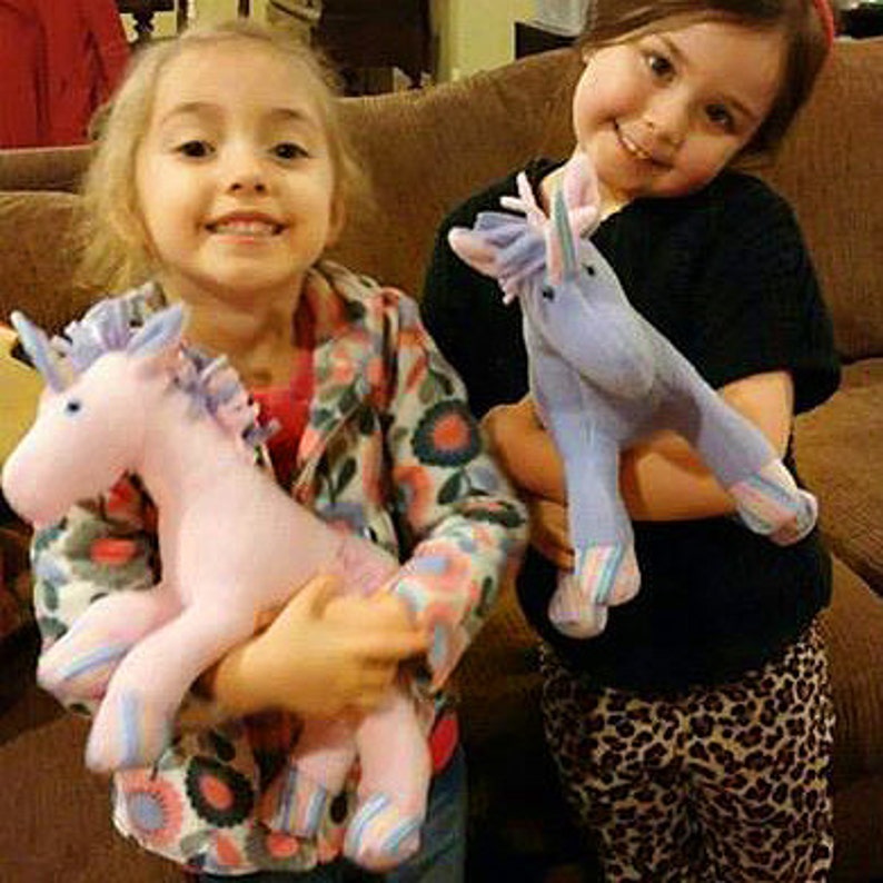 May include: Two young girls are holding plush unicorn toys. One unicorn is pink with blue stripes and the other is purple with blue stripes.