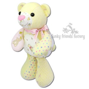 Pode incluir: Um urso de peluche amarelo e rosa com um laço rosa. O urso tem um padrão xadrez e é feito de materiais reciclados.