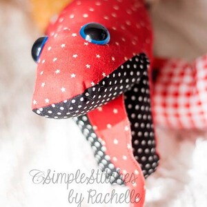 May include: A red and black fabric toy with white polka dots and stars. The toy has blue eyes and a long tail. The toy is on a white surface. Simple Stitches by Rachelle.