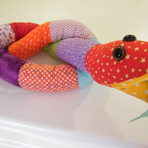 May include: A colorful patchwork snake toy with a red head, yellow and white polka dot belly, and a green tongue. The snake is made of different fabrics with various patterns, including stars, polka dots, and floral designs.