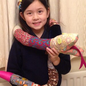 May include: A young girl wearing a navy blue sweater and a headband with colorful buttons holds a large, colorful, patchwork snake toy. The snake is made of different fabrics and has a pink tail.