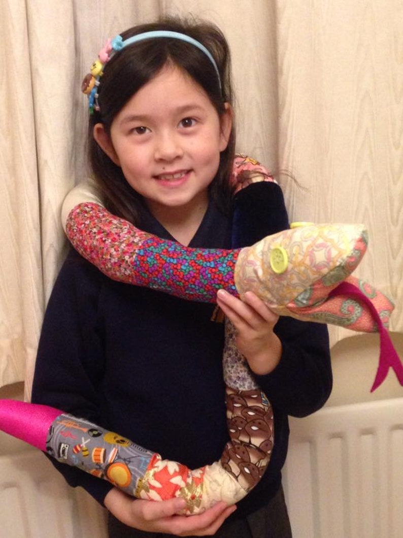 May include: A young girl wearing a navy blue sweater and a headband with colorful buttons holds a large, colorful, patchwork snake toy. The snake is made of different fabrics and has a pink tail.