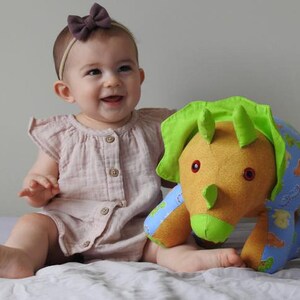 May include: A baby wearing a pink dress and a brown bow headband is playing with a plush green and blue triceratops toy.