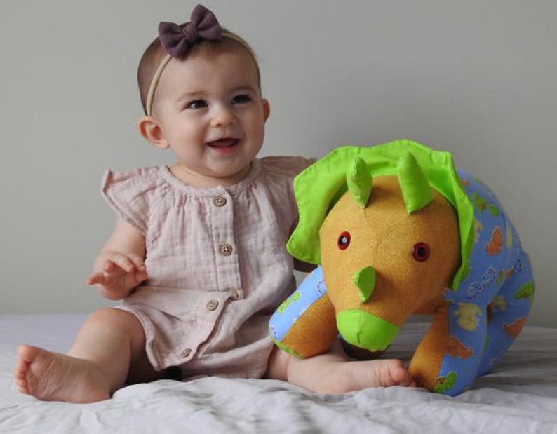 May include: A baby wearing a pink dress and a brown bow headband is playing with a plush green and blue triceratops toy.