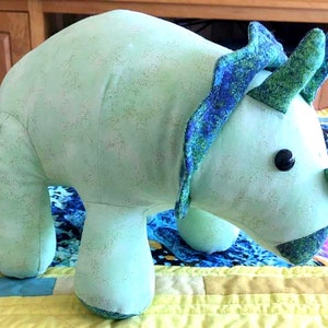 May include: A light blue plush toy shaped like a triceratops dinosaur. The toy has a green and blue patterned frill and a green nose.