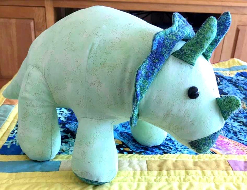 May include: A light blue plush toy shaped like a triceratops dinosaur. The toy has a green and blue patterned frill and a green nose.