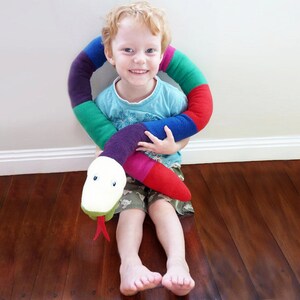 May include: A young child is sitting on a wooden floor and hugging a colorful, striped, fabric snake toy. The snake has a white, plush head with black eyes and a red tongue.