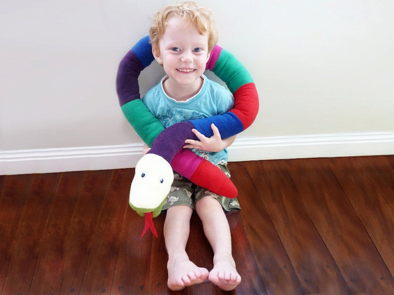 May include: A young child is sitting on a wooden floor and hugging a colorful, striped, fabric snake toy. The snake has a white, plush head with black eyes and a red tongue.