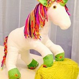 May include: A white stuffed unicorn toy with a rainbow mane and a green horn. The unicorn is standing on a yellow ball of yarn.