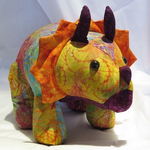 May include: A colourful, handmade stuffed triceratops toy with a yellow, orange and purple patterned fabric. The toy has large, black eyes and a purple nose.