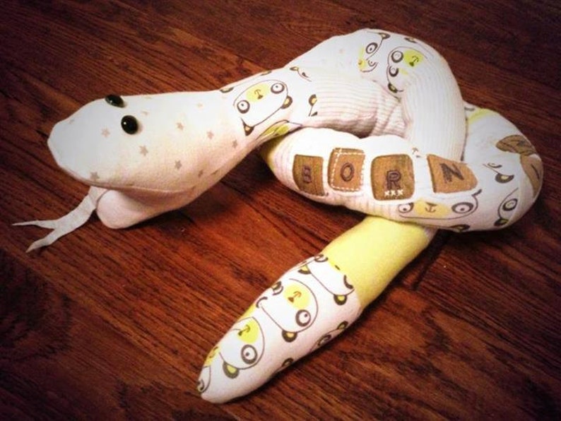 May include: A white and yellow fabric snake toy with black eyes and the word "BORN" spelled out on the body.