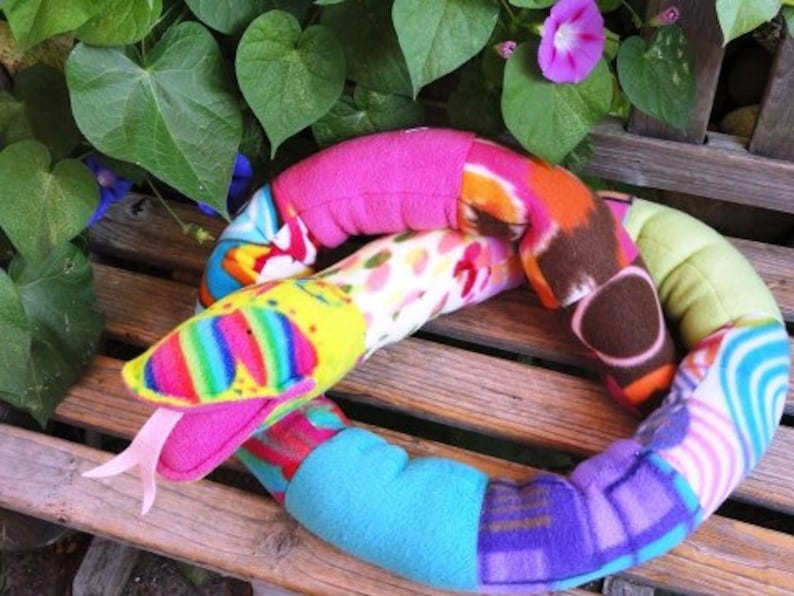 May include: A colorful patchwork snake toy made of different fabrics, including pink, blue, green, brown, and white. The snake has a large, open mouth and a rainbow-colored tongue.