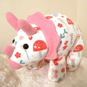 May include: A pink and white plush triceratops toy with a floral and mushroom pattern. The toy has a pink ruffle around the neck and a white background with pink, green, and orange accents.