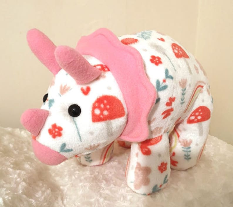 May include: A pink and white plush triceratops toy with a floral and mushroom pattern. The toy has a pink ruffle around the neck and a white background with pink, green, and orange accents.