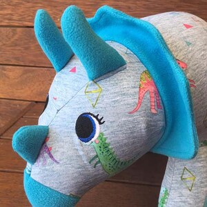 May include: A blue and grey plush toy shaped like a triceratops with a large eye and a green dinosaur printed on the side. The toy has a blue trim around the head and horns.