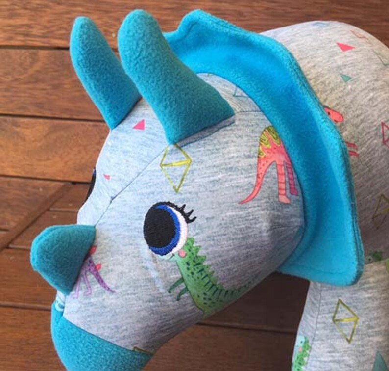 May include: A blue and grey plush toy shaped like a triceratops with a large eye and a green dinosaur printed on the side. The toy has a blue trim around the head and horns.