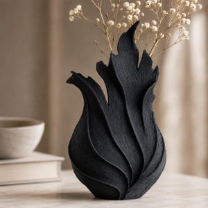 May include: A matte black vase with a textured, organic surface featuring vertical, flame-like ridges that curve upward. The vase has a tapered base and an irregular, pointed rim. Dried white baby's breath flowers are arranged inside the vessel.