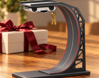 May include: A black magnetic key holder shaped like a curved race track ramp with a white toy car attached to the underside. The structure features red accent lines along the track and a support frame. Two gold-coloured keys hang from the car.