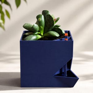 May include: A dark blue square planter with a succulent plant inside. The planter features a triangular side reservoir with a vertical blue tube and a small orange indicator piece at the top, designed for self-watering functionality.