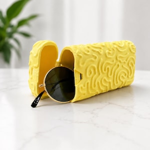 May include: A bright yellow, rectangular, open-ended glasses case with a raised, swirling, brain-like texture on the exterior. A pair of round, dark-tinted sunglasses with thin gold-colored metal frames is partially tucked inside the case.