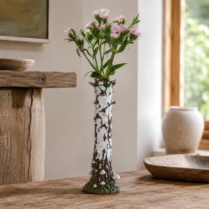 May include: A tall, slender vase designed to resemble a white birch tree trunk with textured bark, dark brown markings, and small white mushroom accents at the base. The vase holds a bouquet of delicate light pink flowers with green stems.