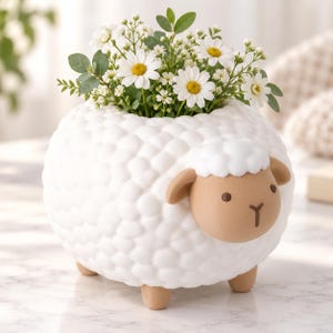 May include: A white ceramic sheep-shaped planter with a textured, bumpy body and a smooth tan face. The sheep has small tan ears and legs, and a tuft of white wool on its head. It is filled with a bouquet of white daisies and green foliage.