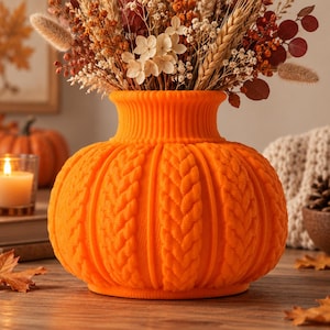 May include: A bright orange vase with a textured, cable-knit sweater pattern and a rounded, pumpkin-like shape. The vase features a wide, ribbed neck and is filled with a dried floral arrangement containing wheat, white flowers, and orange foliage.