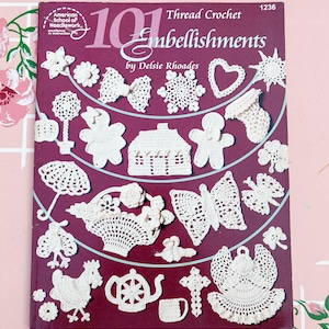 May include: A book cover titled "101 Thread Crochet Embellishments" by Delsie Rhoades. The cover is burgundy with white crochet designs of various shapes, including flowers, a house, a butterfly, and a heart. The American School of Needlework logo is in the upper left corner.