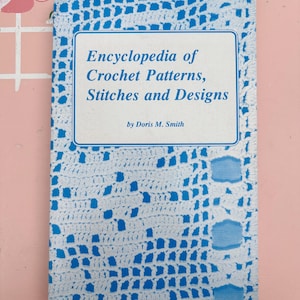 Op de afbeelding: Een boek getiteld "Encyclopedia of Crochet Patterns, Stitches and Designs" door Doris M. Smith. De omslag is wit met een blauw haakpatroon. De titel staat in blauwe tekst in een witte rechthoek.