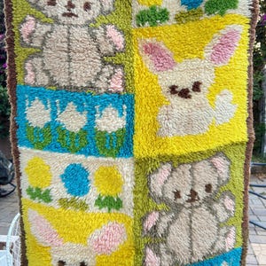 Pode incluir: Uma tapeçaria retangular de tufting com um padrão de grade que alterna painéis de ursinhos de peluche bege, coelhos brancos e motivos florais coloridos em tons de amarelo, azul e verde, contornada por uma borda de fio castanho escuro.
