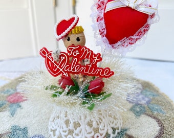 May include: A vintage Valentine's Day decoration featuring a small doll with a red heart hat, a red heart with white lace, and a red sign that says "To My Valentine". The decoration is surrounded by white tinsel and red flowers.