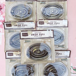 May include: Several antique finish drop pulls from Sears, each in its original packaging. The oval pulls have a floral design in the center. The packaging includes the text "DROP PULL" and "ANTIQUE FINISH".