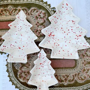 Puede incluir: Tres platos de servir de cerámica blanca con forma de árbol de Navidad con glaseado moteado rojo. Los platos están dispuestos en orden decreciente de tamaño.