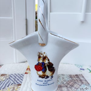 May include: White ceramic basket with a handle, featuring gold trim and a Peter Rabbit and Benjamin Bunny illustration. The basket has a flared shape and a handle with gold accents. The text "Peter Rabbit and Benjamin Bunny" is printed below the illustration.